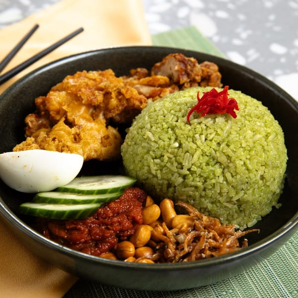 A black bowl of green rice, fried chicken, cucumber, egg, sambal, peanuts, and anchovies.
