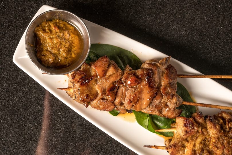 Grilled chicken skewers with a side of peanut sauce on a white rectangular plate.