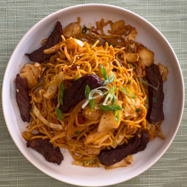 A white bowl filled with noodles, beef strips, cabbage, and green onions on a green placemat.