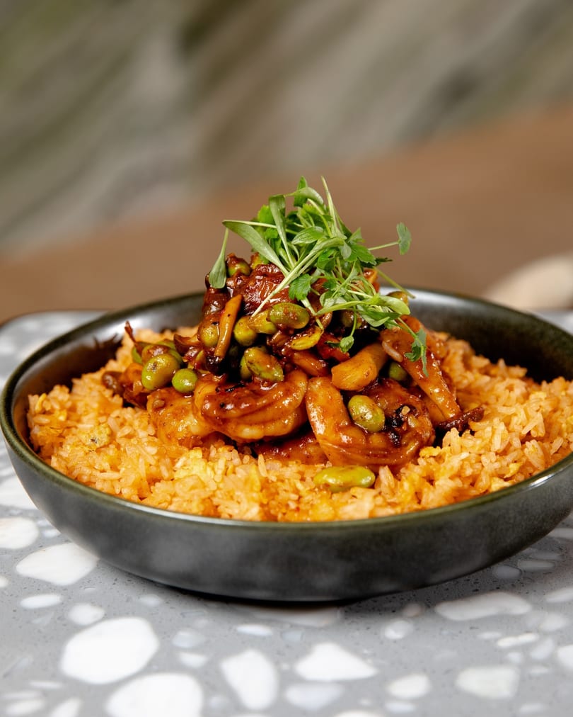 Shrimp and edamame stir-fry over orange rice, garnished with fresh greens in a dark bowl.
